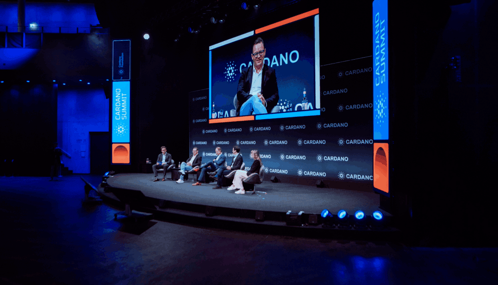 Four people sit on a stage during a panel discussion at the Cardano Summit. Large digital screens behind them display the Cardano Summit name and a close-up of one panelist speaking, all under blue, professional lighting.